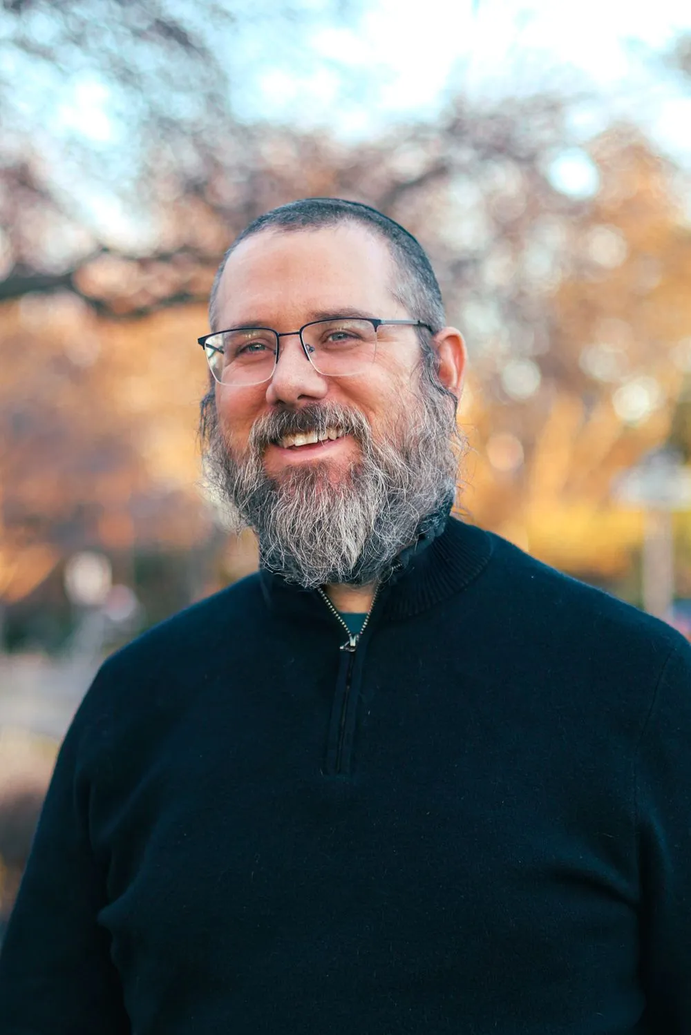 Aaron Silverman smiling outdoors in autumn, wearing glasses and a dark sweater, warm trees behind him.