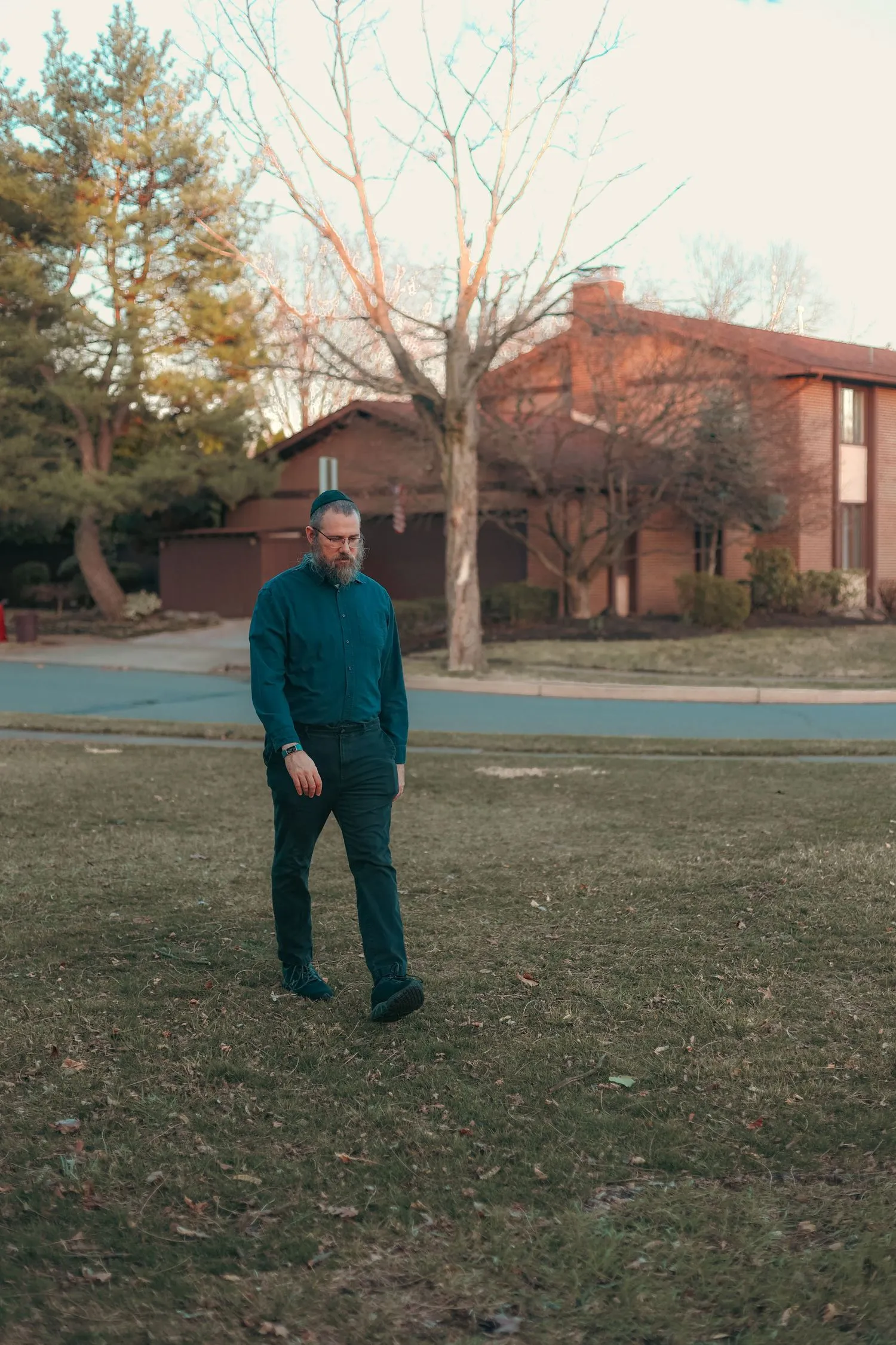 Aaron Silverman walking across a lawn at dusk in a teal shirt, a bare tree and brick houses behind him